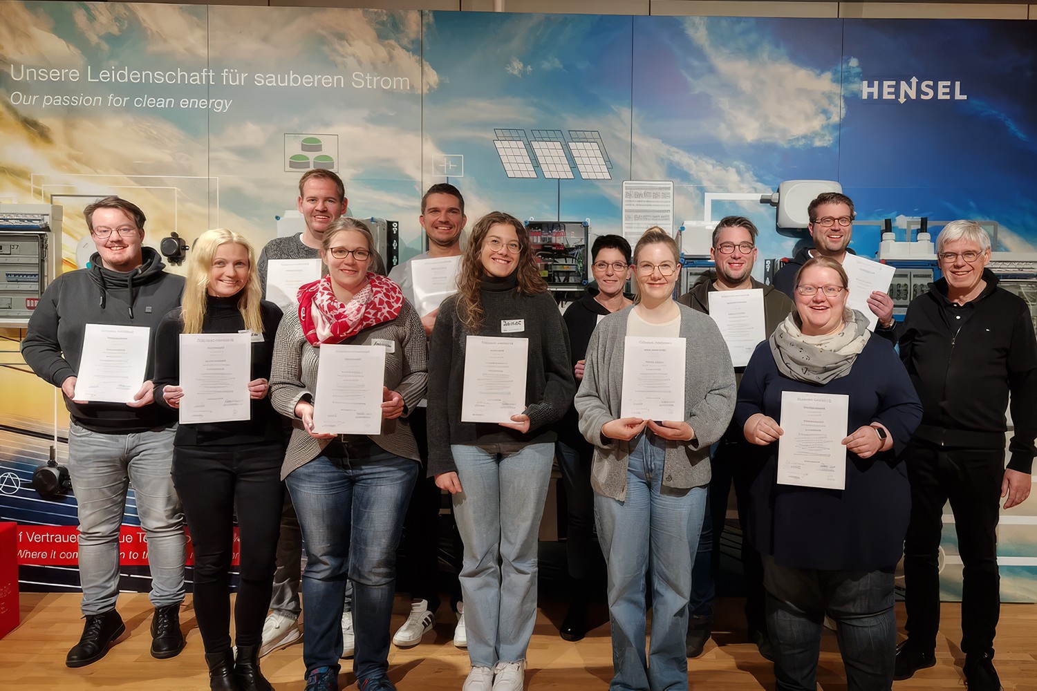 Gruppe von 13 frisch zertifizierten AGILean Mastern bei Hensel Electric, alle halten ihre Zertifikate in die Kamera, vor einem Hensel-Markenbanner im Hintergrund